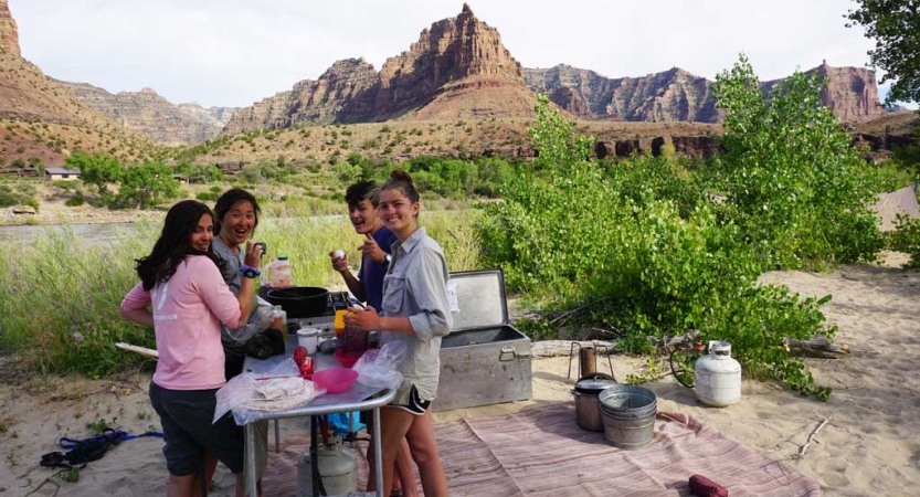 A group of students prepare food on an outdoor table with mountains in the background. 